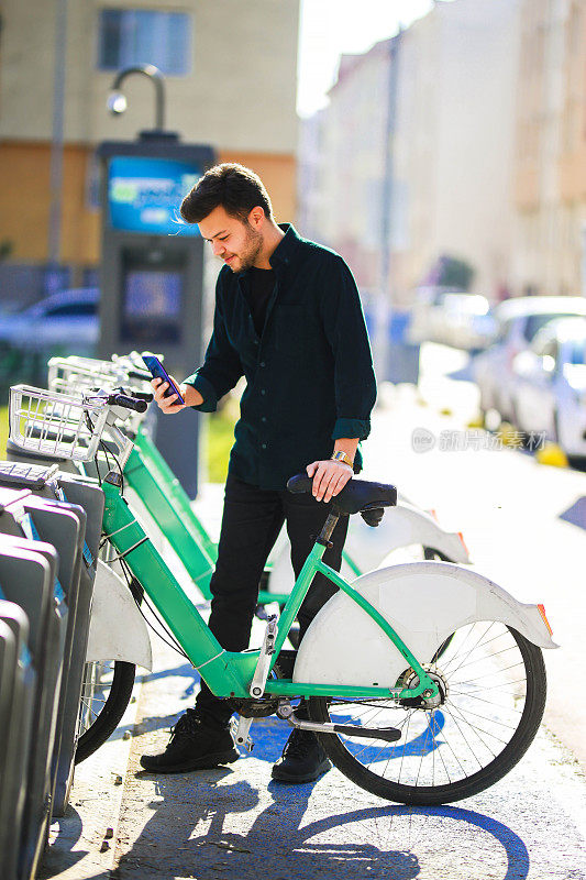 Young brown-haired man renting from bike rental station with his mobile phone. city ​​bike transportation concept. young man renting a bike in the city